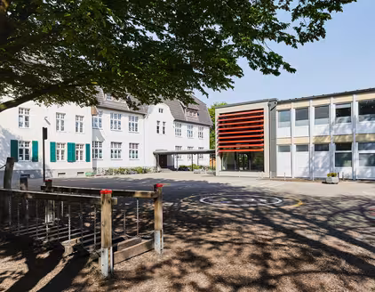 Schule mit roten Lamellen an Fensterfront und Spielplatz vorne links