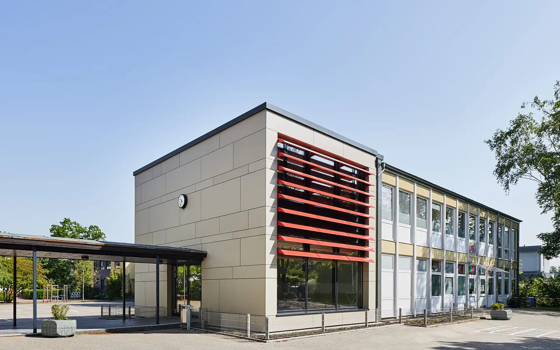 Schule mit roten Lamellen an Fensterfront, überdachter Weg zu Nebengebäude