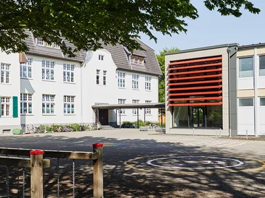 Schule mit roten Lamellen an Fensterfront und Spielplatz vorne links